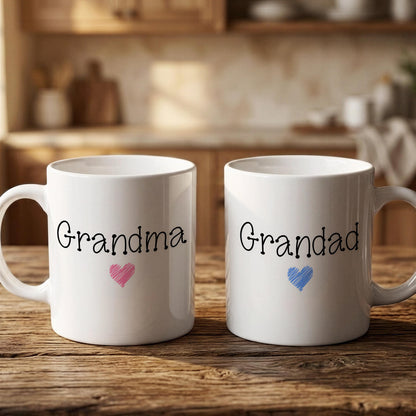 Two white mugs with the text "Grandma" and "Grandpa" printed on them, along with a heart symbol, are placed on a wooden surface.