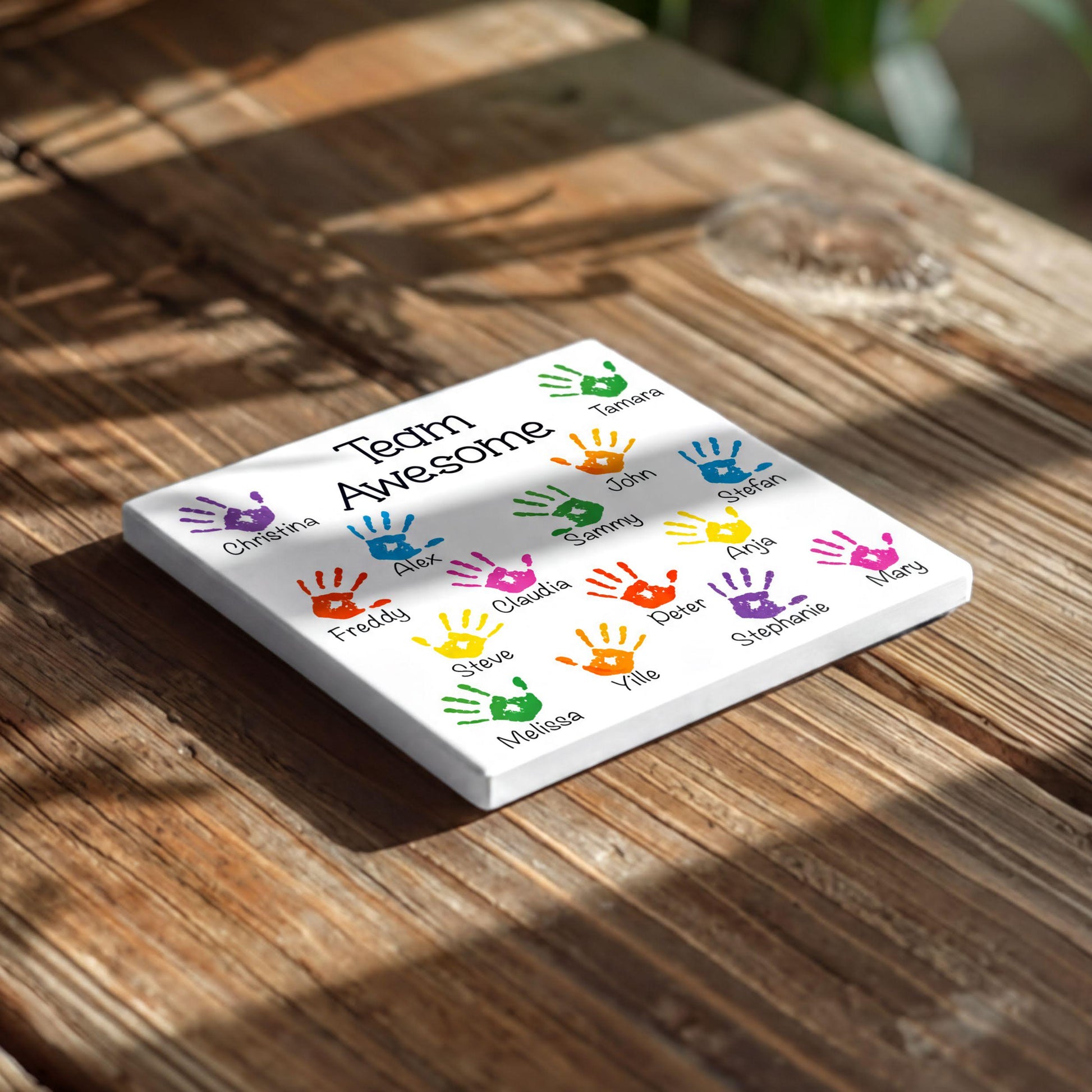 A white square tile with colorful handprint designs on it, placed on a wooden surface.