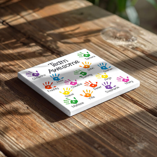 A white square tile with colorful handprint designs on it, placed on a wooden surface.