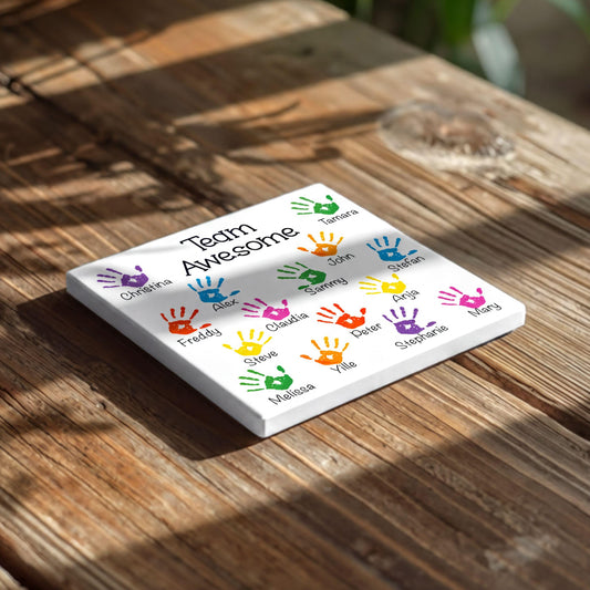 A white square tile with colorful handprint designs on it, placed on a wooden surface.