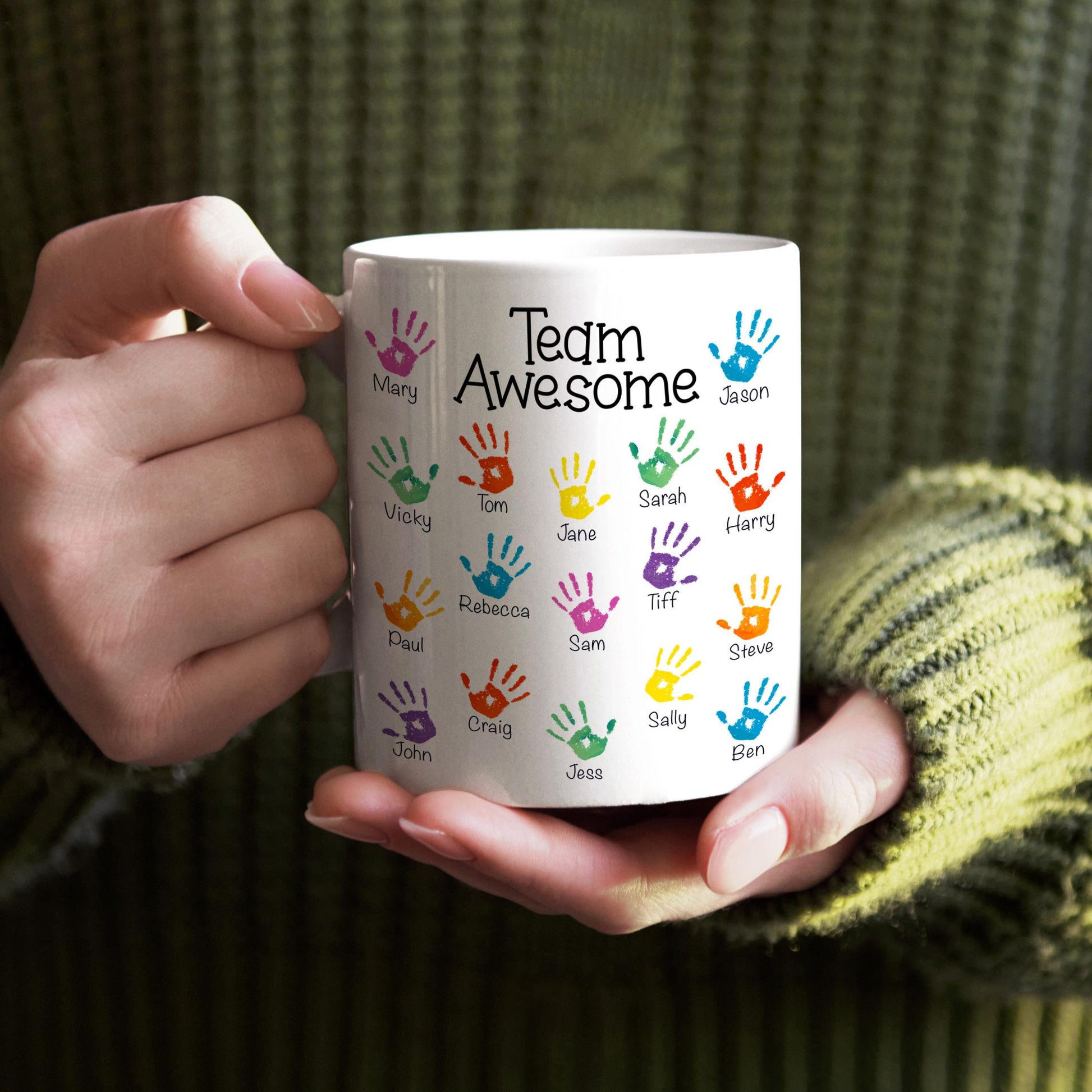 A person is holding a white mug with colorful handprints on it, and the mug has the text "Team Awesome" printed on it.