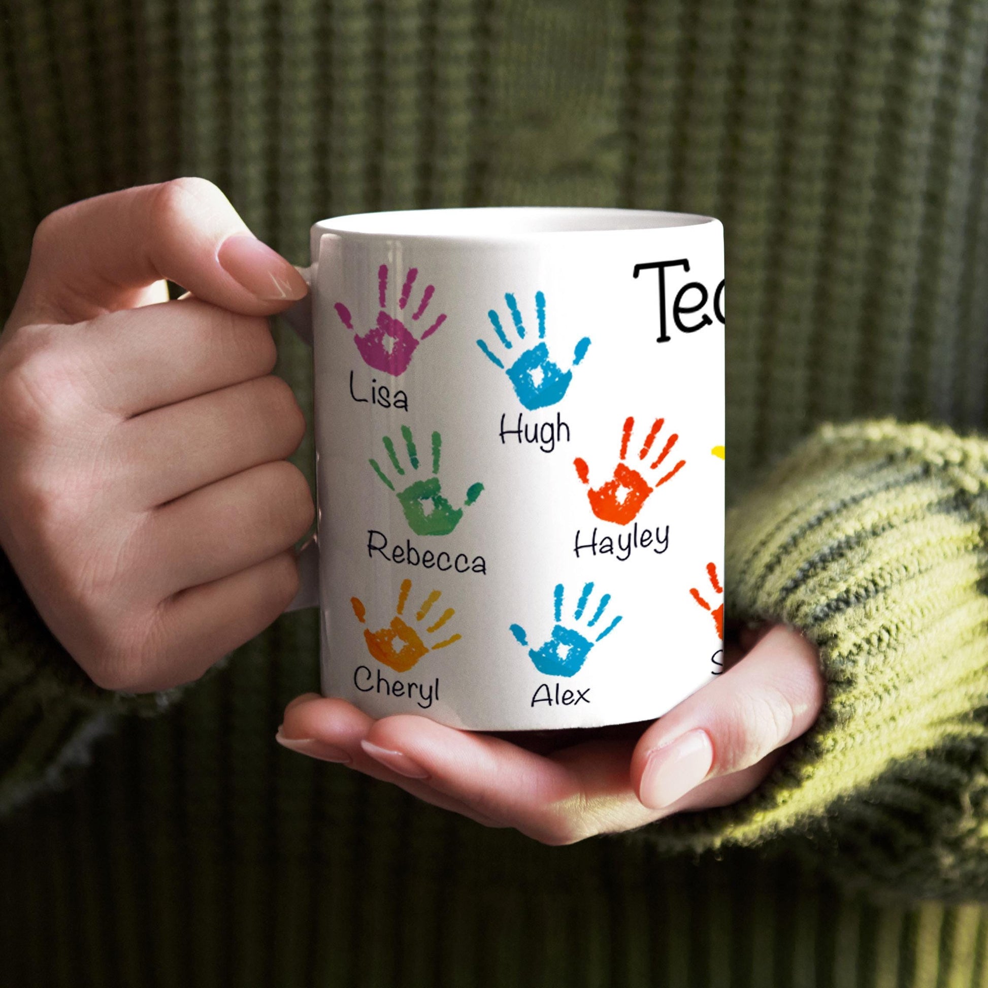 A person is holding a white mug with colorful handprints on it, each containing a name.