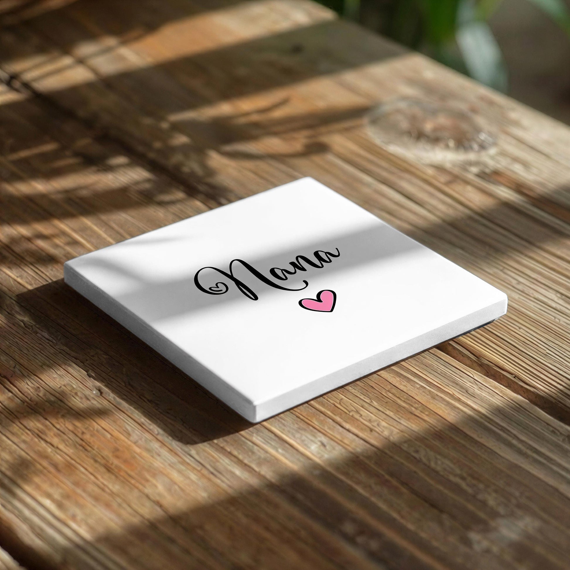 A white square card with the word "Nana" written on it, along with a heart symbol, placed on a wooden surface.