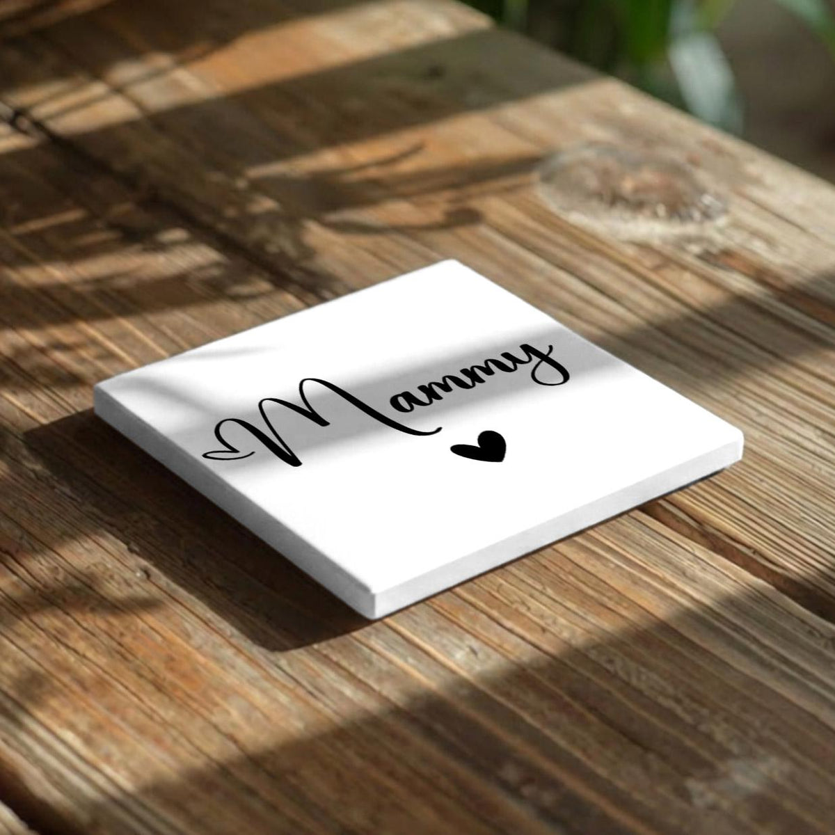 A white square coaster with the word "Mammy" written on it, along with a heart symbol, placed on a wooden surface.