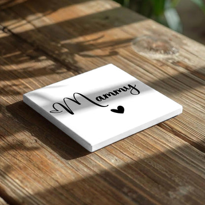 A white square coaster with the word "Mammy" written on it, along with a heart symbol, placed on a wooden surface.