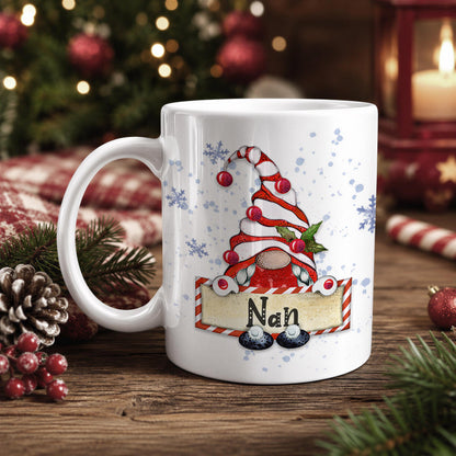 A white mug with a Christmas-themed design featuring a gnome wearing a red and white striped hat, surrounded by snowflakes and pine branches.