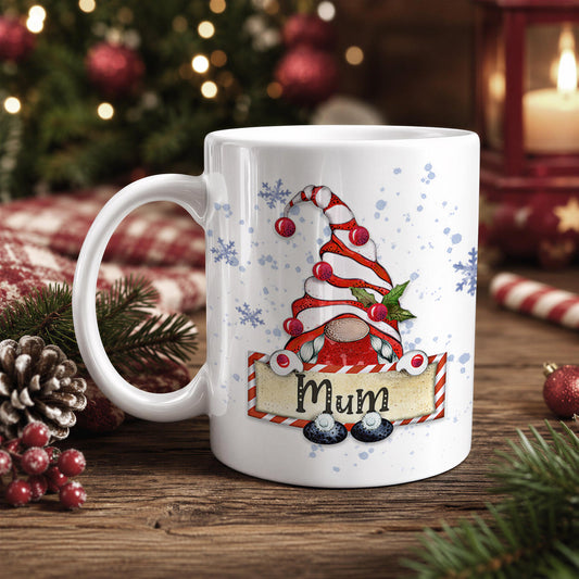 A white mug with a Christmas-themed design featuring a gnome wearing a red and white striped hat, surrounded by snowflakes and a banner that says "Mum". The mug is placed on a wooden surface, with Christmas decorations and ornaments visible in the background.