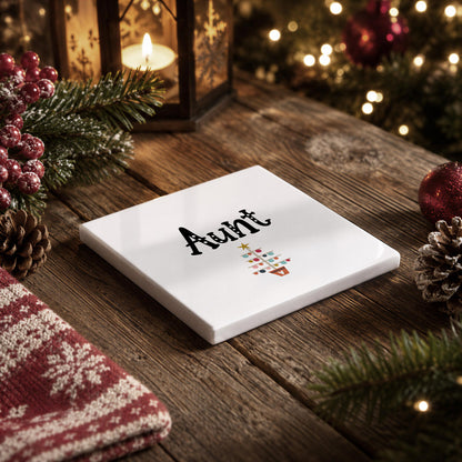 A white square coaster with the word "Aunt" written on it, surrounded by Christmas decorations such as pine cones, a lantern, and a tree.
