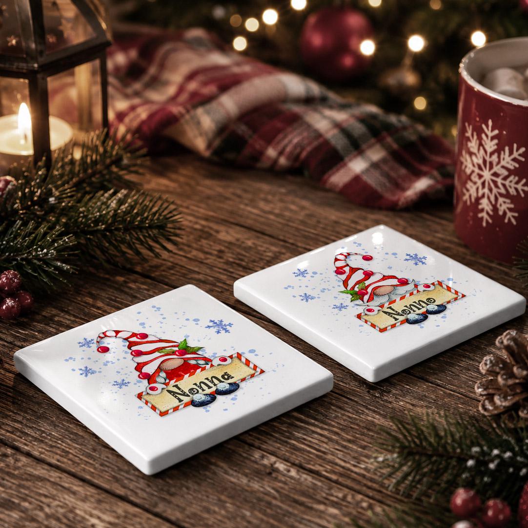 two white coasters with a gnome design, placed on a wooden surface. The coasters are decorated with snowflakes and the name "Nonna". There is also a red mug and a lantern in the background, adding to the festive atmosphere.