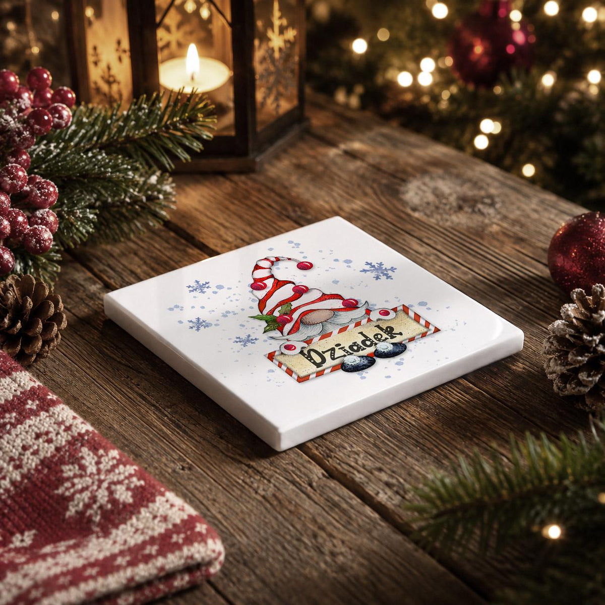 A white square coaster with a gnome design sits on a wooden table, surrounded by Christmas decorations such as pine cones, berries, and a lit lantern.