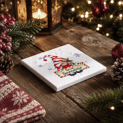 A white coaster with a Christmas-themed design sits on a wooden table, surrounded by holiday decorations such as pine cones, berries, and a lit lantern.