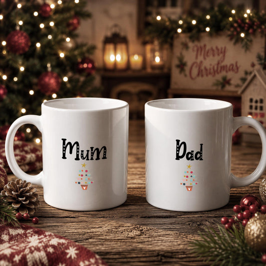 Two white mugs with the names "Mum" and "Dad" printed on them, placed on a wooden surface with Christmas decorations in the background.