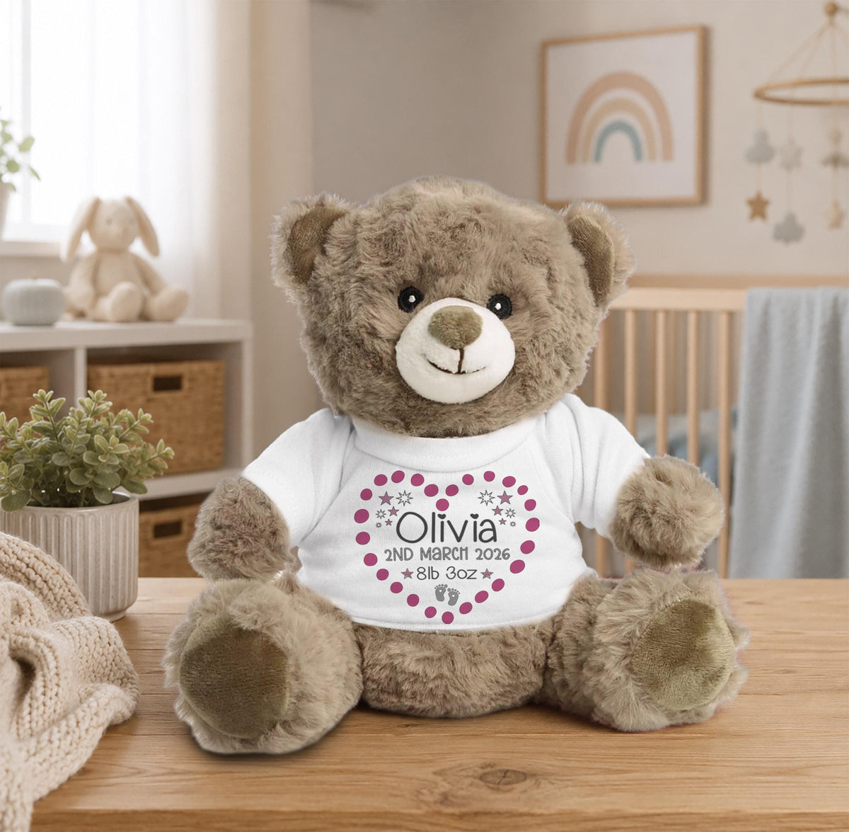 A brown teddy bear wearing a white t-shirt with a heart design sits on a wooden table in a room with a window and a plant.