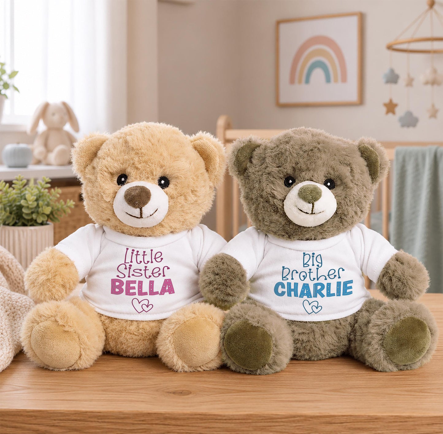 Two teddy bears, one brown and one gray, sit on a wooden surface. The brown teddy bear is wearing a white shirt with the text "Little Sister Bella" and the gray teddy bear is wearing a blue shirt with the text "Big Brother Charlie".