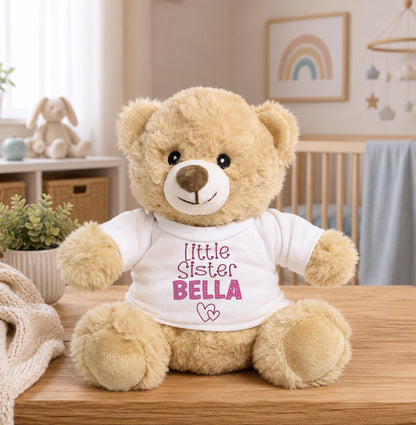 A brown teddy bear wearing a white shirt with the text "Little Sister Bella" sits on a wooden table.