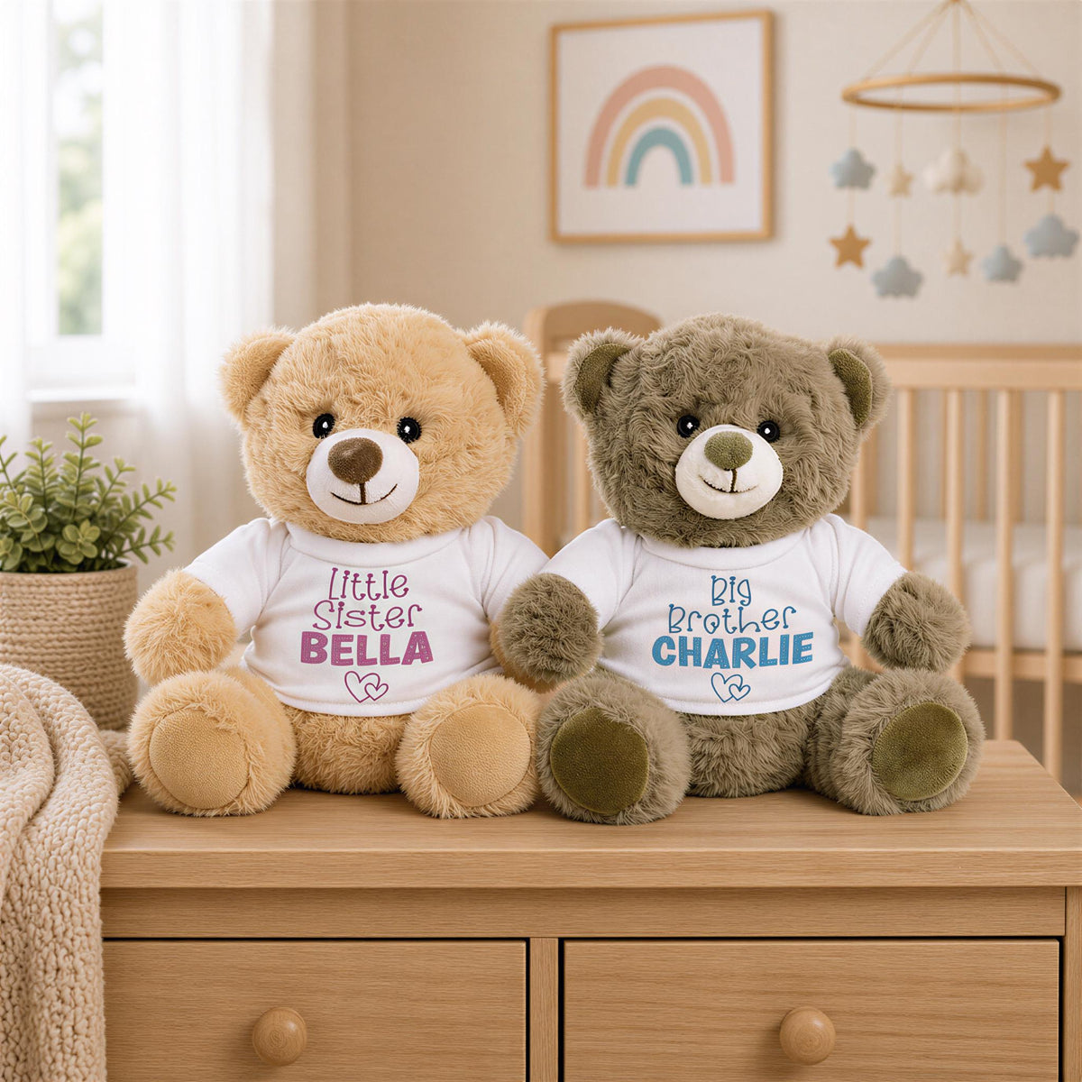 Two teddy bears, one light brown and one dark brown, sit on a wooden dresser in a nursery. The teddy bears are wearing white shirts with the names "Little Sister Bella" and "Big Brother Charlie" printed on them.