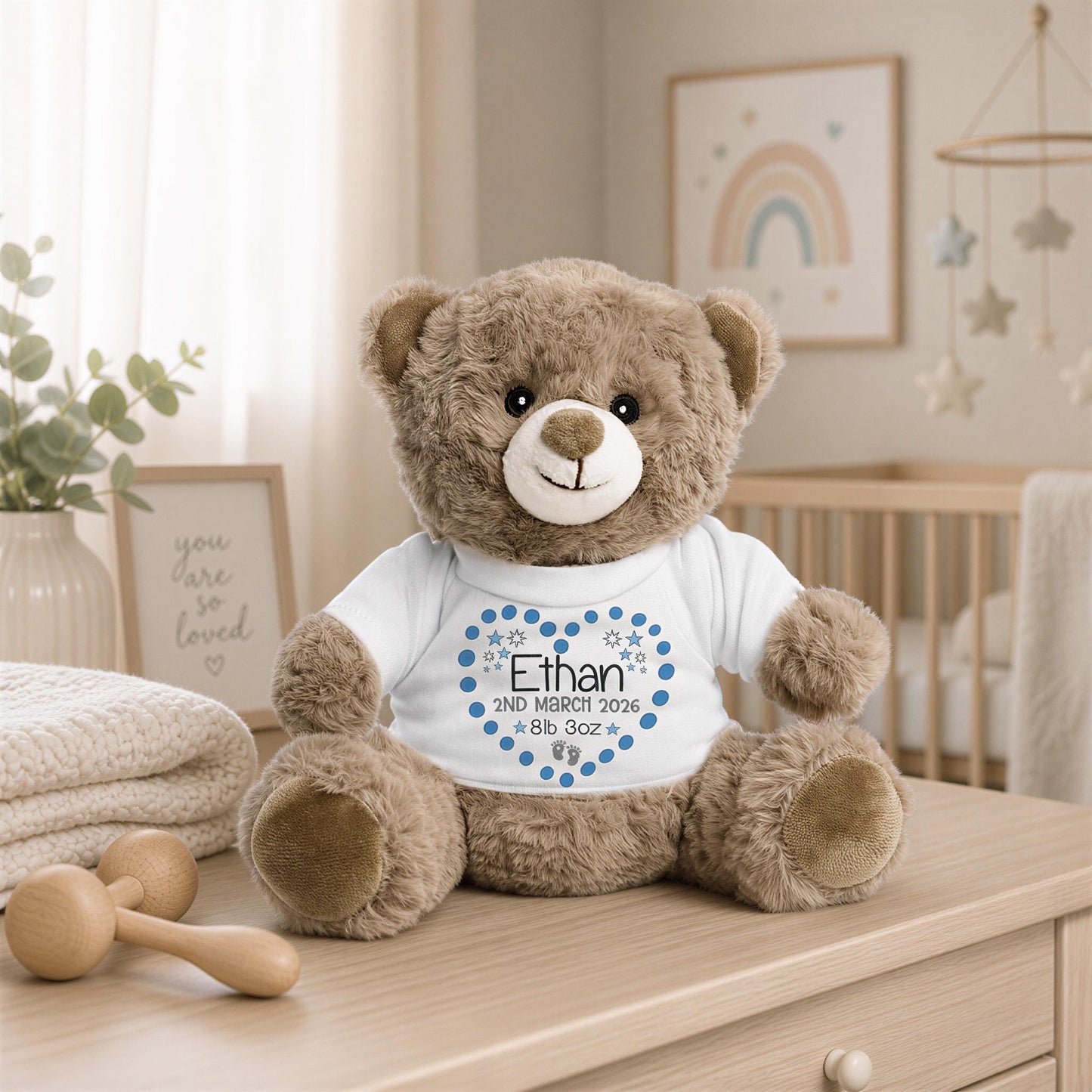 A brown teddy bear wearing a white shirt with a blue heart and the name "Ethan" printed on it, sitting on a wooden dresser in a nursery setting.