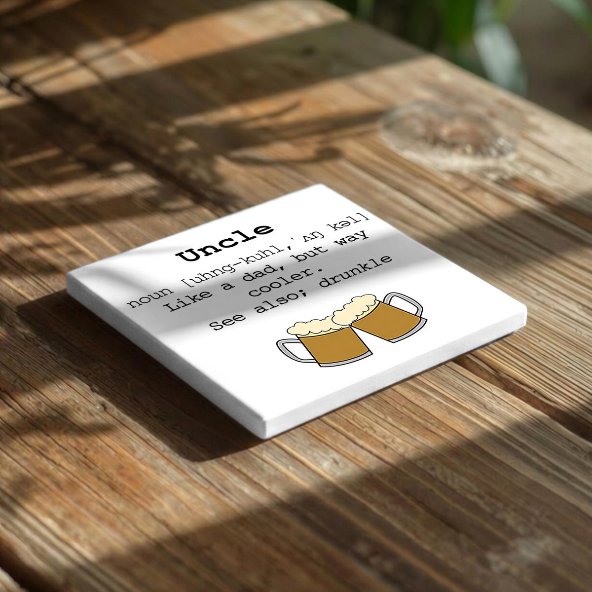 a white square coaster with a humorous definition of "Uncle" written on it, placed on a wooden surface.