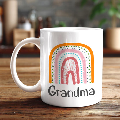 A white ceramic mug with a colorful rainbow design and the word "Grandma" printed on it, placed on a wooden surface.