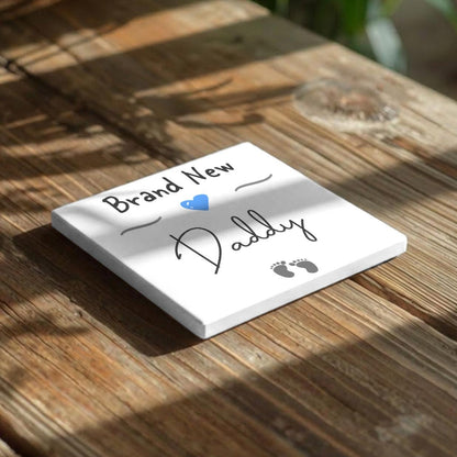 A white square tile with the text "Brand New Daddy" written on it, along with a blue heart symbol, placed on a wooden surface.