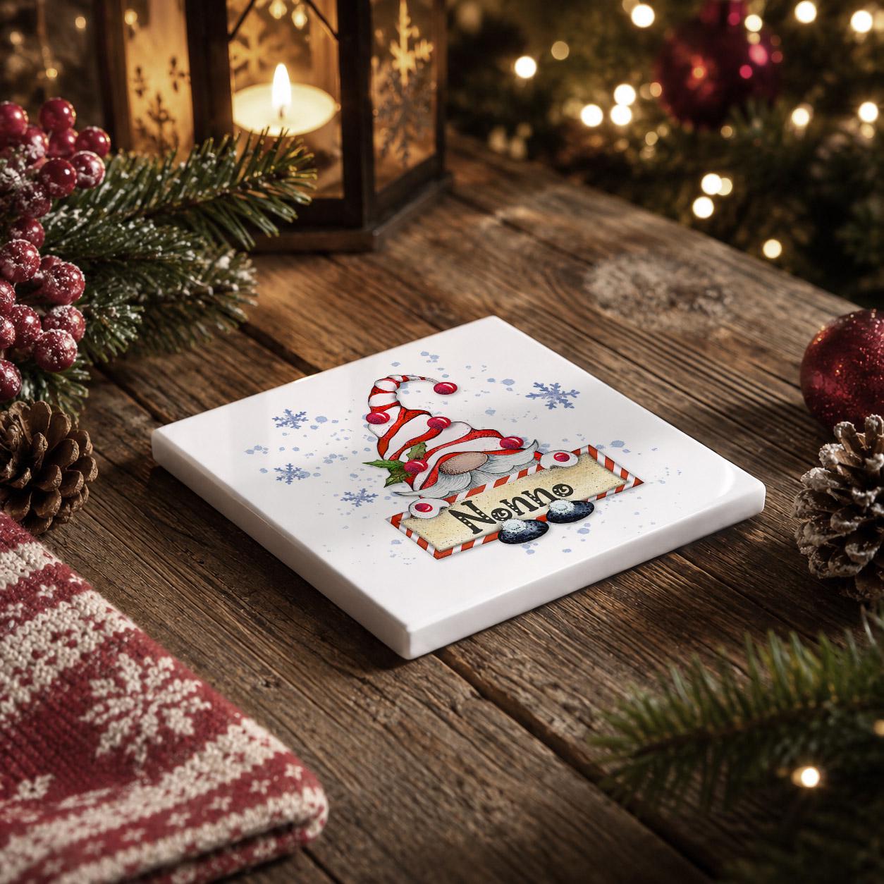 A white square coaster with a red and white striped gnome hat design sits on a wooden surface, surrounded by Christmas decorations such as pine cones, berries, and a lit lantern.
