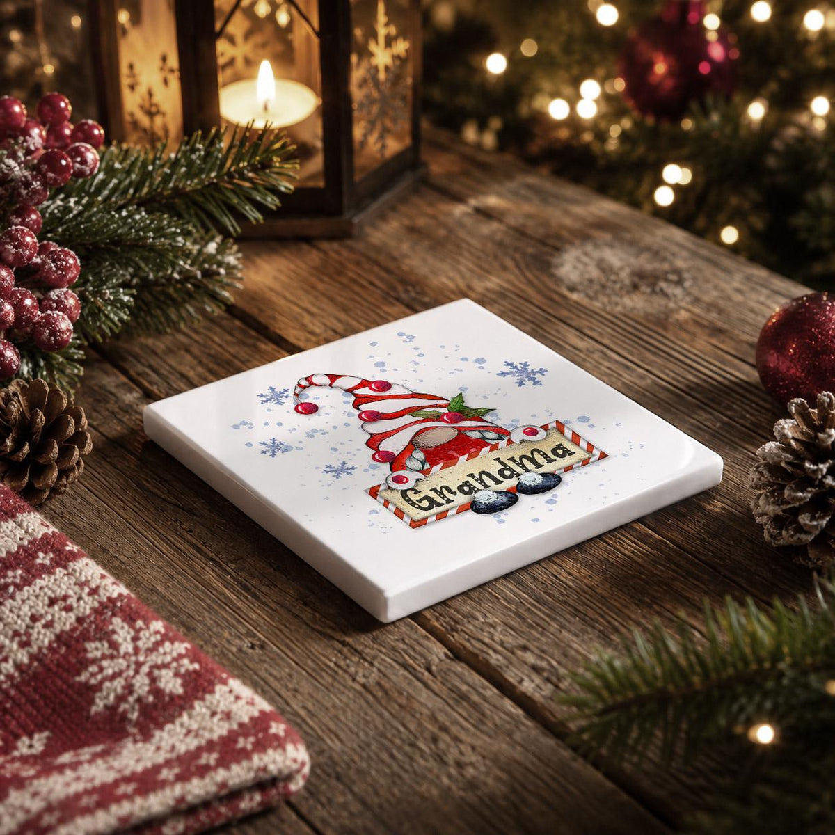 A white square coaster with a Christmas-themed design sits on a wooden table, surrounded by holiday decorations such as pine cones, berries, and a lit lantern.