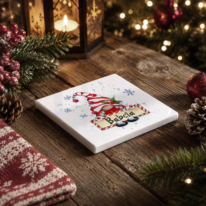 A white square coaster with a Christmas-themed illustration of a gnome is placed on a wooden surface, surrounded by holiday decorations such as pine cones, berries, and a lit lantern.