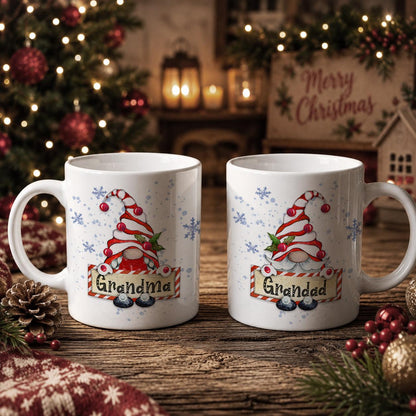 Two white coffee mugs with Christmas-themed designs, featuring gnomes and snowflakes, are placed on a wooden surface. The mugs are decorated with the text "Grandma" and "Grandad" printed on them. In the background, there is a Christmas tree and other holiday decorations, creating a festive atmosphere.