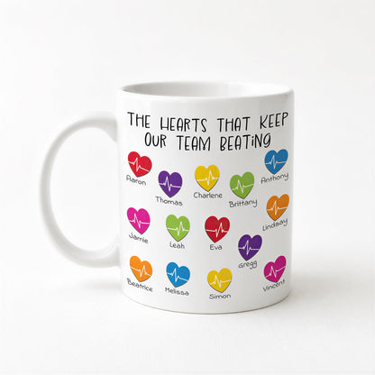 A white ceramic mug with a printed design featuring colorful heart icons and text that reads "The hearts that keep our team beating".