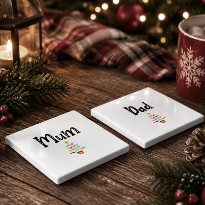 two white coasters with the names "Mum" and "Dad" printed on them, placed on a wooden surface with a Christmas tree and other holiday decorations in the background.