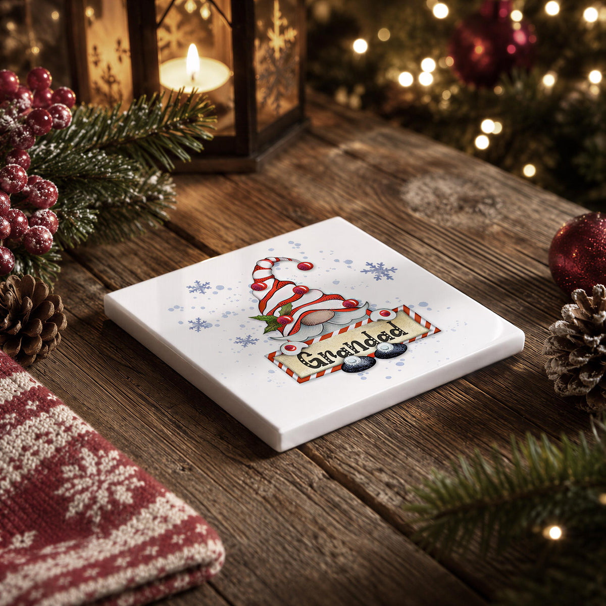 A white square coaster with a Christmas-themed illustration and the word "Grandad" written on it, placed on a wooden surface with pine cones, a red ornament, and a lantern in the background.