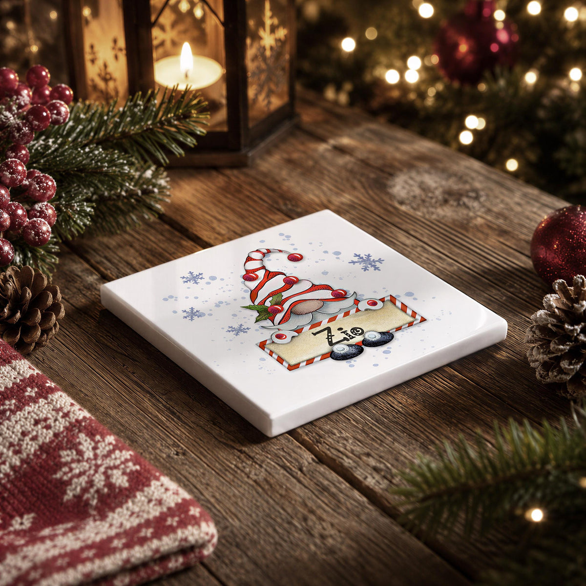 A white square coaster with a Christmas-themed illustration of a gnome wearing a red and white striped hat, surrounded by snowflakes and pine cones, sits on a wooden surface.