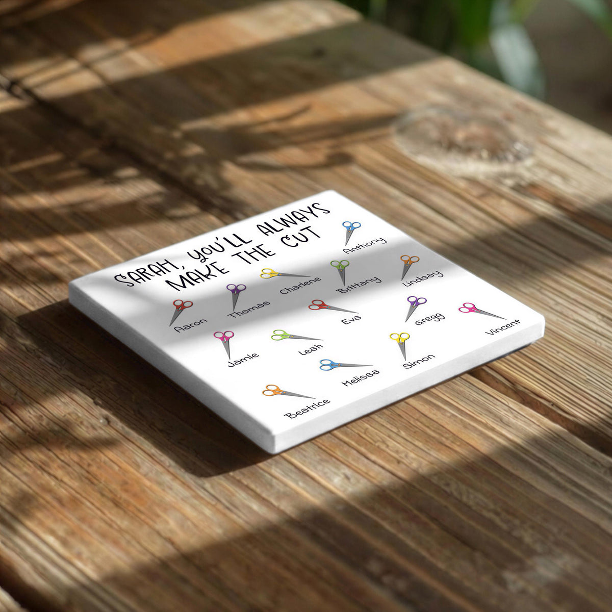 A white square coaster with a list of names and scissors printed on it, placed on a wooden surface.