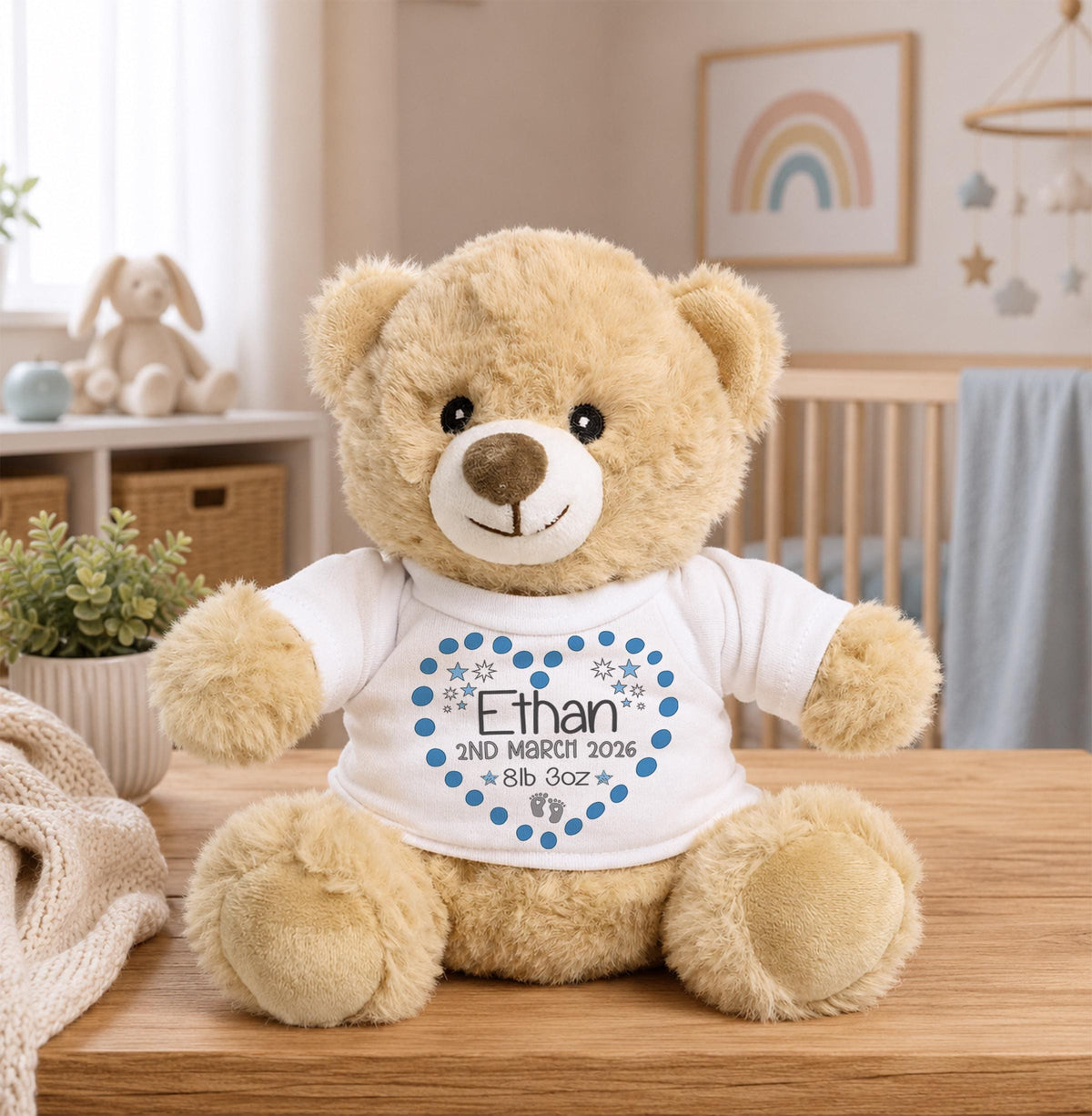 A brown teddy bear wearing a white shirt with a blue heart and the name "Ethan" printed on it, sitting on a wooden table in a nursery setting.