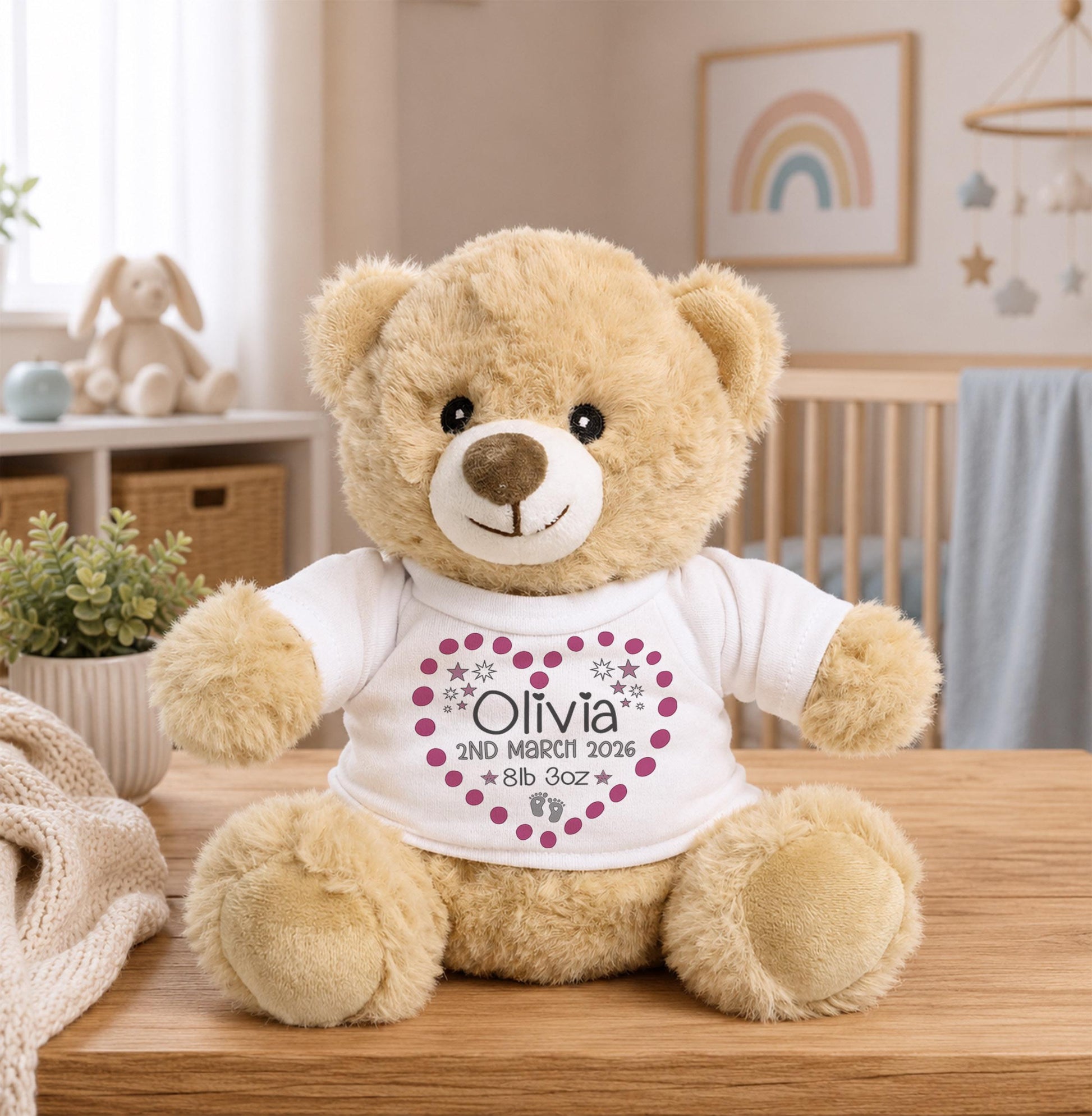 A brown teddy bear wearing a white shirt with a heart and the text "Olivia" and "2nd Month 2016" printed on it, sitting on a wooden table in a nursery.