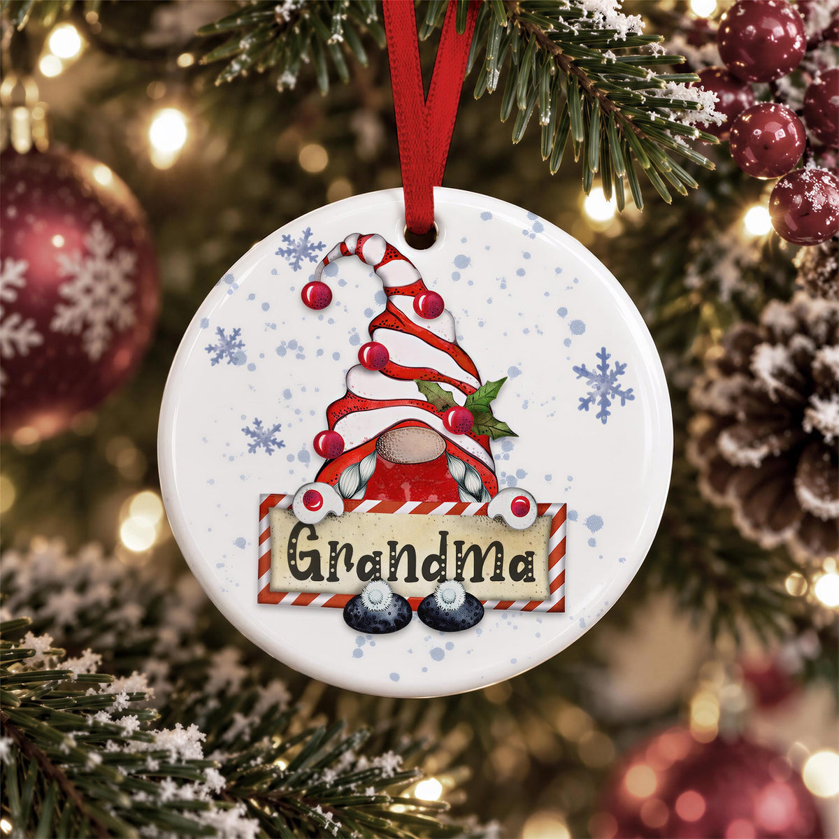 A white ornament with a red ribbon and a gnome design hangs on a Christmas tree, surrounded by pine branches and red and gold ornaments.