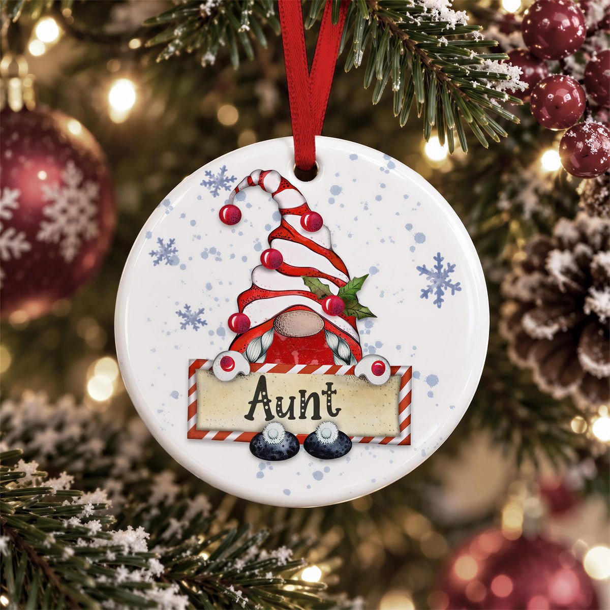 A white ornament with a red ribbon and a gnome design hangs on a Christmas tree, surrounded by snowflakes and ornaments.