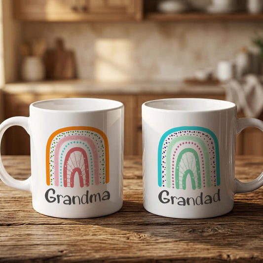 Two white mugs with colorful rainbows and the text "Grandma" and "Grandad" printed on them, placed on a wooden surface.