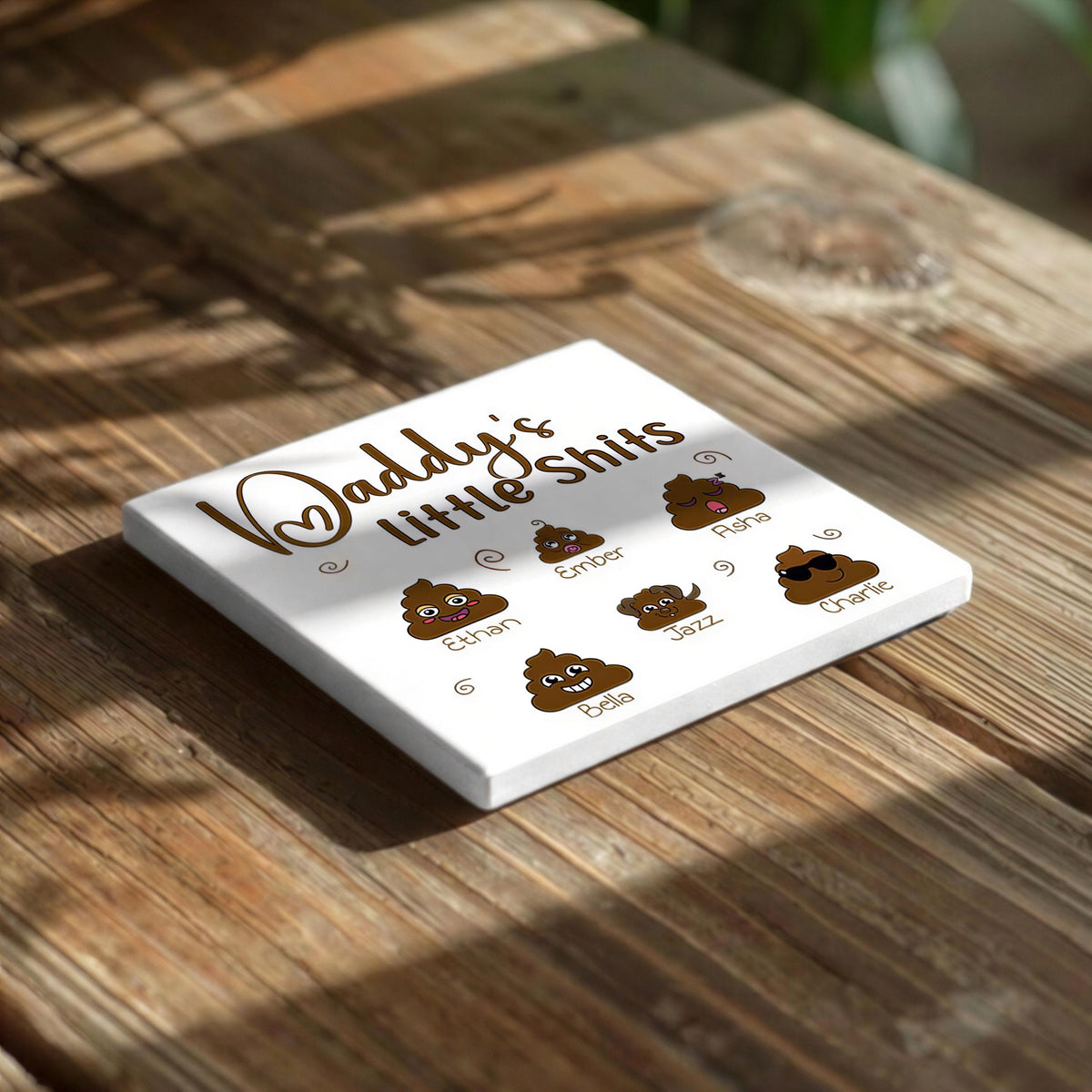 A white coaster with a humorous text and six cartoon images of different types of poop on it, placed on a wooden table.