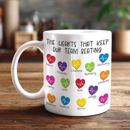 A white mug with a printed message that reads "The hearts that keep our team beating" along with various heart icons representing different people.