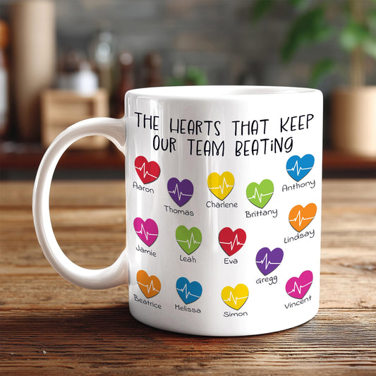 A white mug with a printed message that reads "The hearts that keep our team beating" along with various heart icons representing different people.